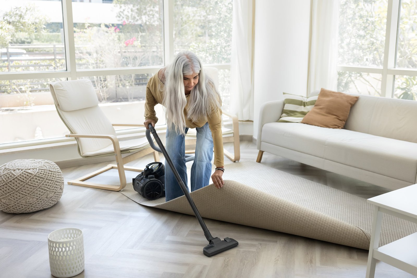 Woman with long grey hair vacuuming under a rug in a bright, modernly furnished living room.
