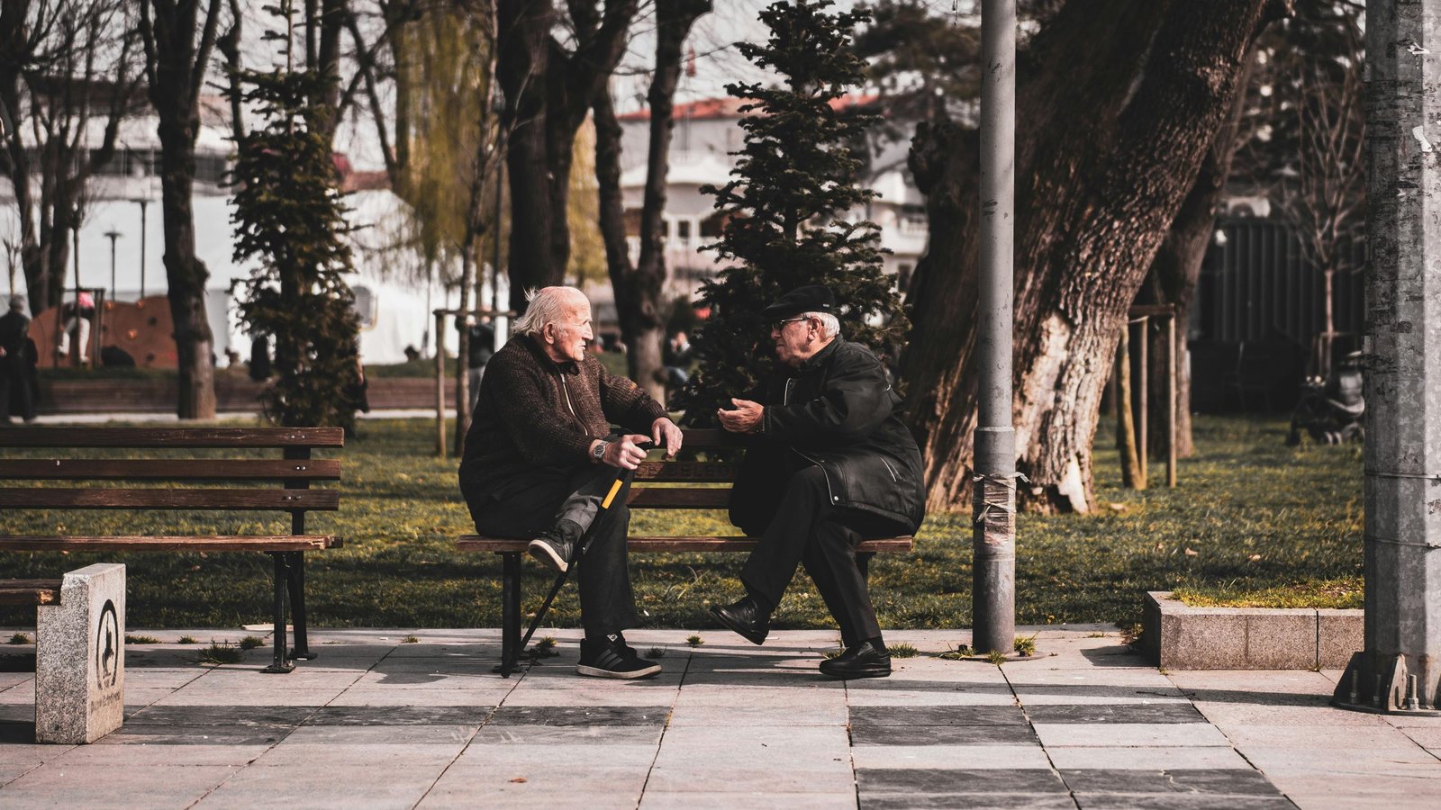 two elderly men wearing coats and hats, sitting on a park bench, talking to each other